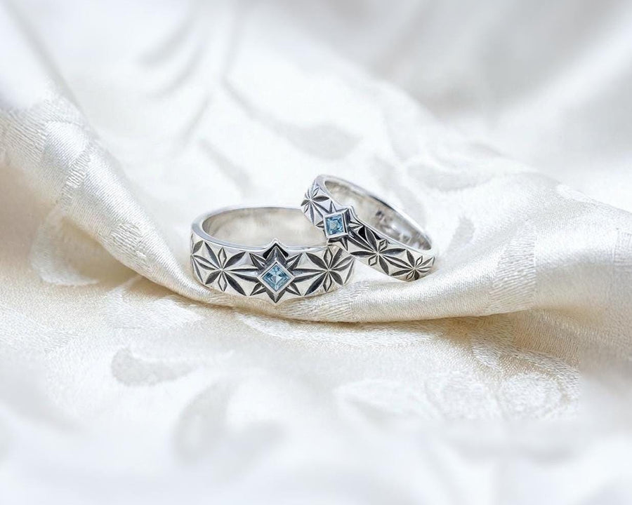 A close-up studio photograph of a matching pair of 925 sterling silver wedding bands resting on a bright, reflective white surface scattered with tiny crystals. The rings feature a continuous, deeply engraved pattern of geometric starbursts or celestial stars with radiating lines. The primary band has a square, princess-cut light blue gemstone, likely blue topaz or aquamarine, bezel-set directly into the center of the prominent front-facing star.