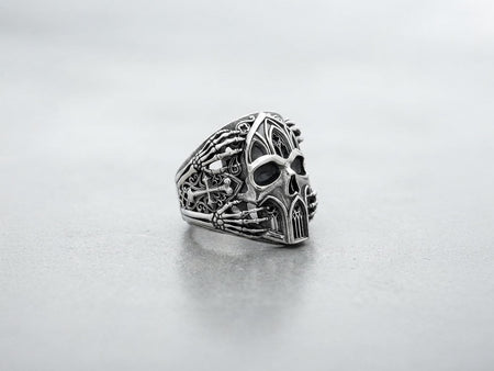 A close-up studio photograph of a 925 sterling silver skull ring on a black background. The skull features gothic cathedral windows carved into its forehead and chin. Skeletal hands grip the sides of the skull. The recessed areas are deeply oxidized to a dark matte gray, contrasting with polished silver highlights.