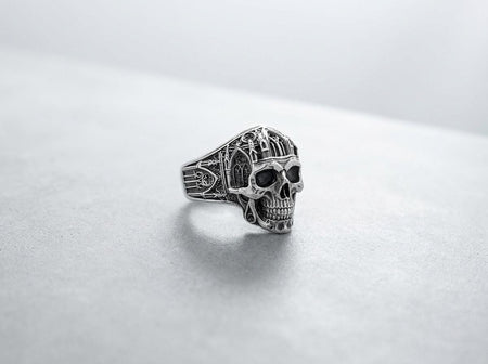 A close-up studio photograph of a 925 sterling silver skull ring resting on a sleek black background. The skull's cranium is intricately sculpted to resemble a gothic cathedral, complete with miniature lancet windows, arched doorways, and tall spires. The recessed areas of the silver are deeply oxidized to a dark matte gray, contrasting sharply with the polished highlights of the bone structure and architectural details.