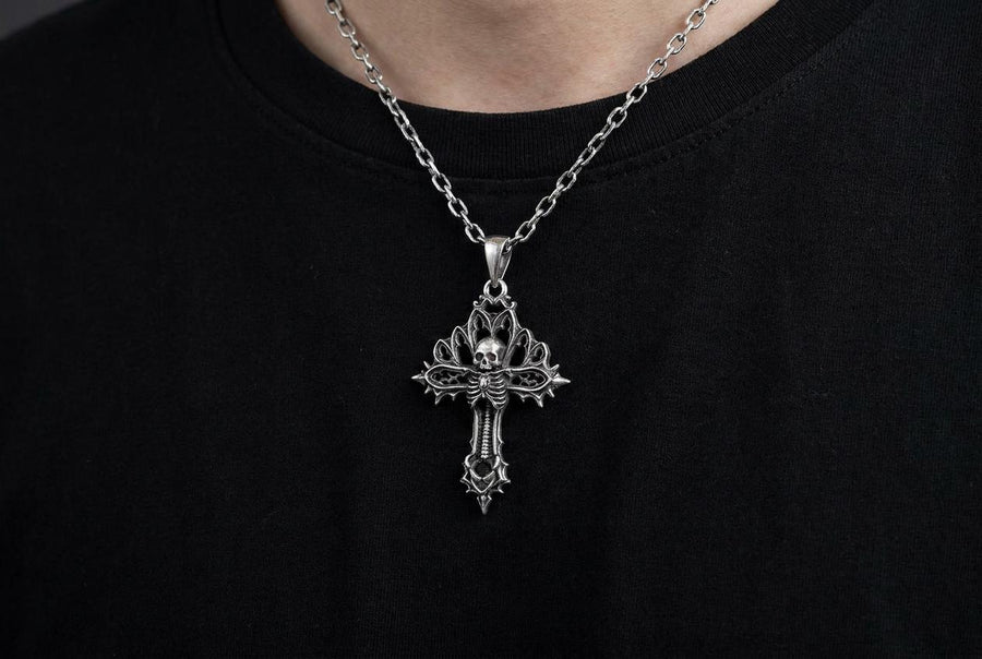 A close-up studio photograph of a 925 sterling silver gothic cross pendant resting on a light, finely textured surface. The core of the cross features a detailed skull, ribcage, and a spine extending down the bottom arm. The upper arms are crafted with openwork medieval tracery resembling cathedral windows. The recessed silver is heavily oxidized to a dark gray, contrasting sharply with the polished bone and architectural highlights.