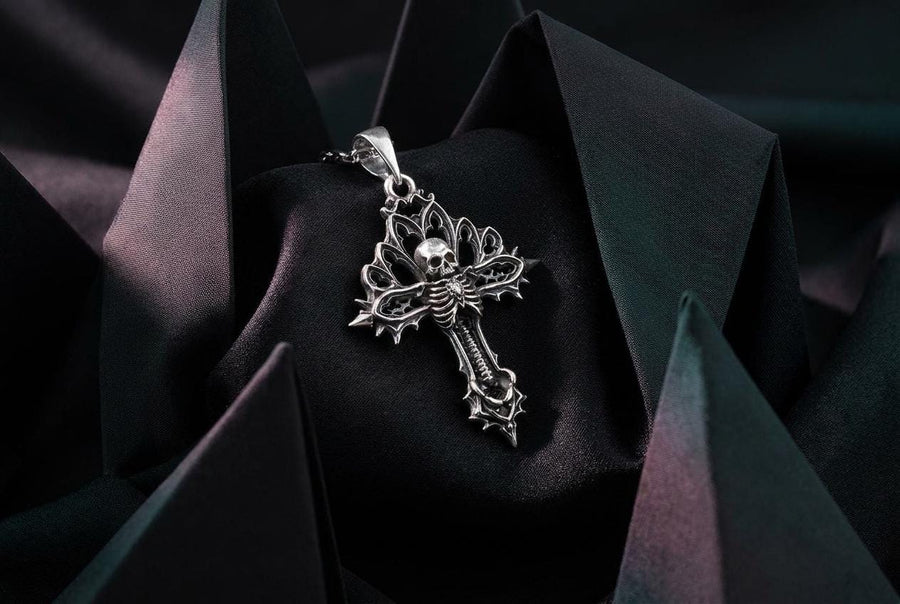 A close-up studio photograph of a 925 sterling silver gothic cross pendant resting on a light, finely textured surface. The core of the cross features a detailed skull, ribcage, and a spine extending down the bottom arm. The upper arms are crafted with openwork medieval tracery resembling cathedral windows. The recessed silver is heavily oxidized to a dark gray, contrasting sharply with the polished bone and architectural highlights.