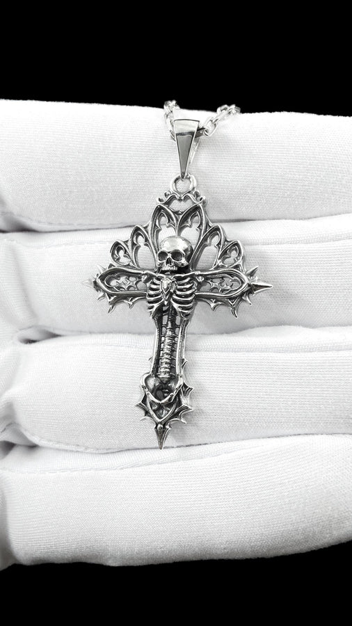 A close-up studio photograph of a 925 sterling silver gothic cross pendant resting on a light, finely textured surface. The core of the cross features a detailed skull, ribcage, and a spine extending down the bottom arm. The upper arms are crafted with openwork medieval tracery resembling cathedral windows. The recessed silver is heavily oxidized to a dark gray, contrasting sharply with the polished bone and architectural highlights.