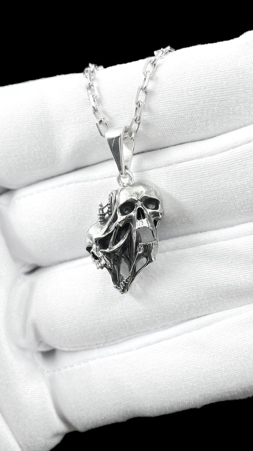 A side-profile view of the silver skull pendant showcases the breathtaking depth of the sculpted elements. The skulls appear stretched and elongated, morphing into each other. Dark oxidation heavily shadows the deeply recessed areas of the bone.