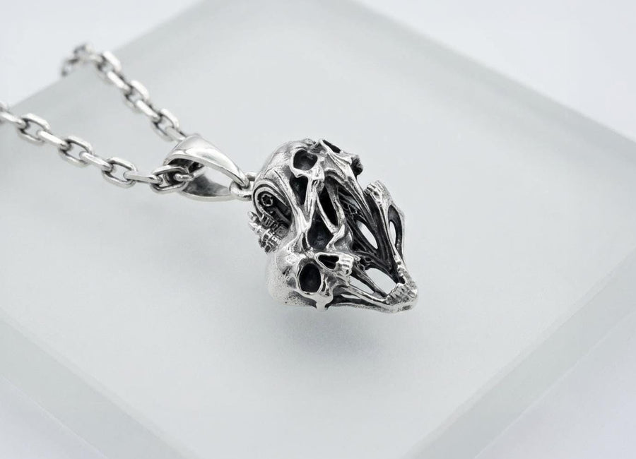 A side-profile view of the silver skull pendant showcases the breathtaking depth of the sculpted elements. The skulls appear stretched and elongated, morphing into each other. Dark oxidation heavily shadows the deeply recessed areas of the bone.