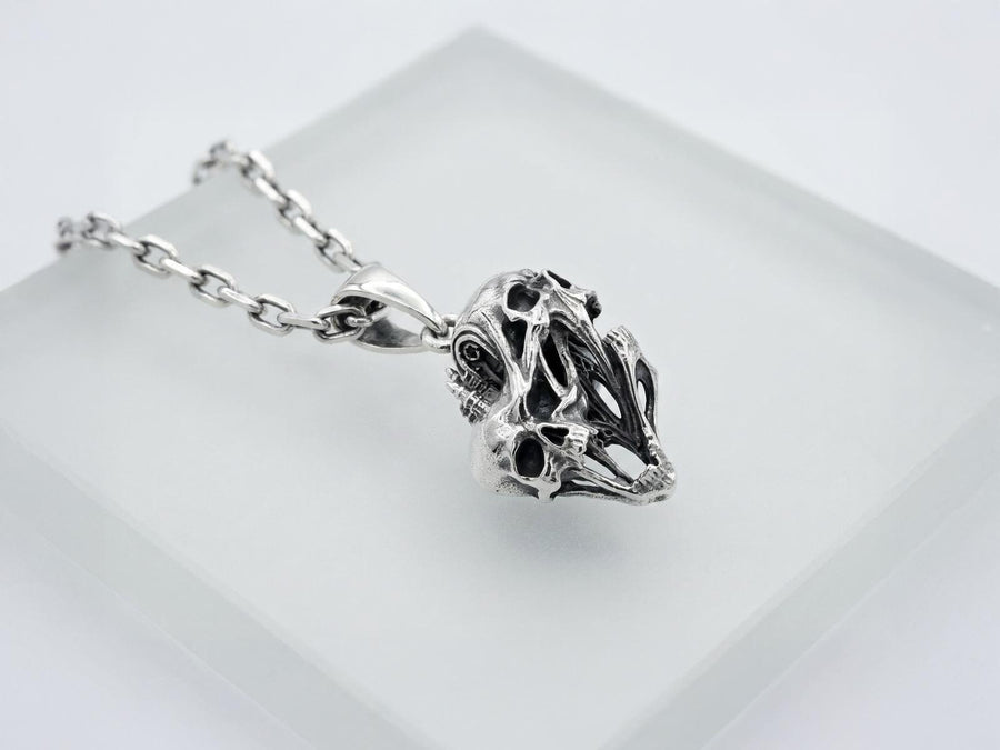 A side-profile view of the silver skull pendant showcases the breathtaking depth of the sculpted elements. The skulls appear stretched and elongated, morphing into each other. Dark oxidation heavily shadows the deeply recessed areas of the bone.