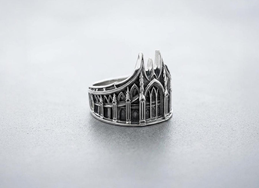 This solid 925 sterling silver gothic cathedral ring features soaring spires and intricately carved pointed arches. Resting on a grey surface, this oxidized silver band showcases detailed medieval tracery and classic stonework columns, perfect for a dark romance aesthetic. A striking 925 sterling silver architecture ring capturing the monumental scale of medieval sanctuaries. Bright lighting illuminates the polished silver gothic spires, contrast