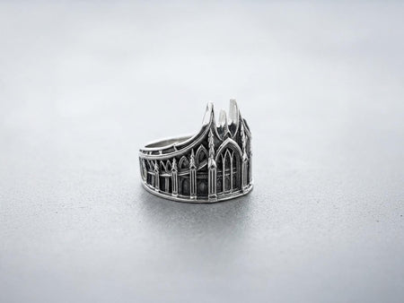 This solid 925 sterling silver gothic cathedral ring features soaring spires and intricately carved pointed arches. Resting on a grey surface, this oxidized silver band showcases detailed medieval tracery and classic stonework columns, perfect for a dark romance aesthetic. A striking 925 sterling silver architecture ring capturing the monumental scale of medieval sanctuaries. Bright lighting illuminates the polished silver gothic spires, contrast