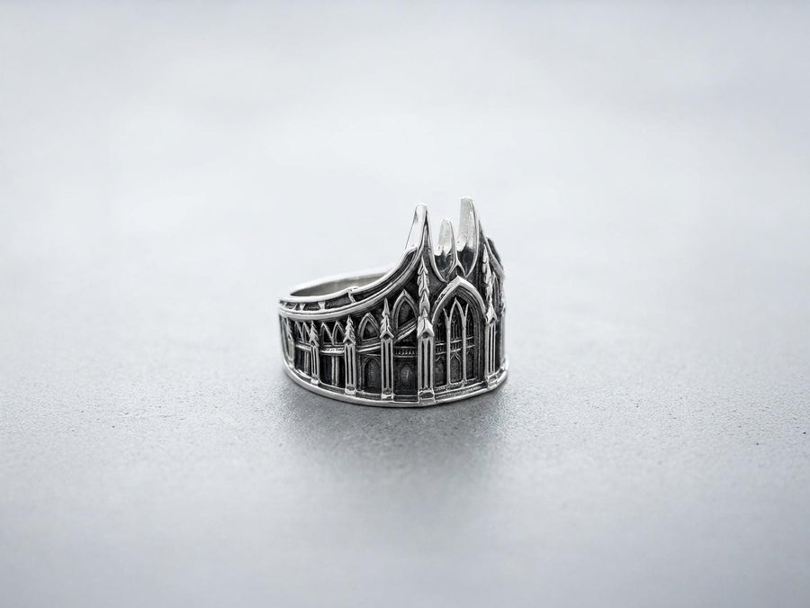 This solid 925 sterling silver gothic cathedral ring features soaring spires and intricately carved pointed arches. Resting on a grey surface, this oxidized silver band showcases detailed medieval tracery and classic stonework columns, perfect for a dark romance aesthetic. A striking 925 sterling silver architecture ring capturing the monumental scale of medieval sanctuaries. Bright lighting illuminates the polished silver gothic spires, contrast