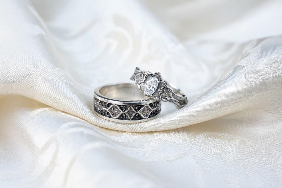 A close-up studio photograph of a matching 925 sterling silver gothic wedding ring set on a grey surface. The wide mens band and the delicate womens engagement ring both feature intricate medieval tracery and carved quatrefoils - highlighted by a dark oxidized background. This striking artisan bridal set perfectly captures the monumental scale of medieval architecture. Bright lighting illuminates the polished silver edges and the sparkling oval g
