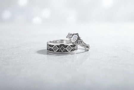 A close-up studio photograph of a matching 925 sterling silver gothic wedding ring set on a grey surface. The wide mens band and the delicate womens engagement ring both feature intricate medieval tracery and carved quatrefoils - highlighted by a dark oxidized background. This striking artisan bridal set perfectly captures the monumental scale of medieval architecture. Bright lighting illuminates the polished silver edges and the sparkling oval g