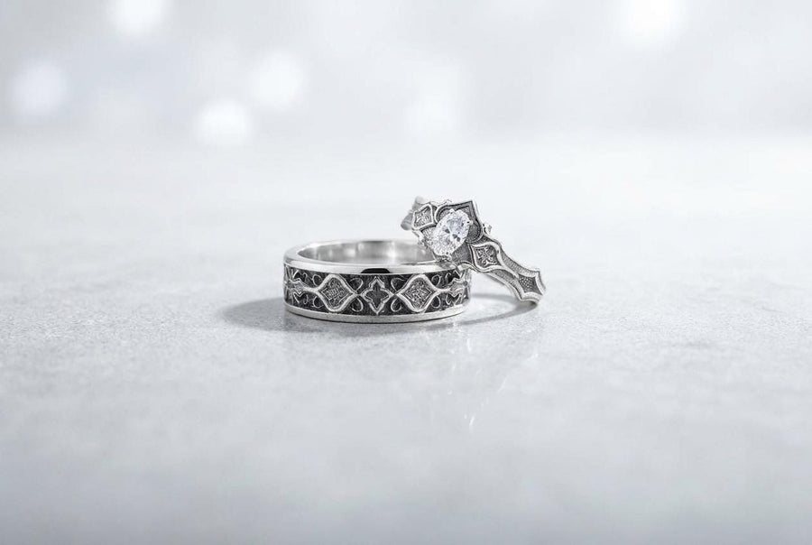 A close-up studio photograph of a matching 925 sterling silver gothic wedding ring set on a grey surface. The wide mens band and the delicate womens engagement ring both feature intricate medieval tracery and carved quatrefoils - highlighted by a dark oxidized background. This striking artisan bridal set perfectly captures the monumental scale of medieval architecture. Bright lighting illuminates the polished silver edges and the sparkling oval g