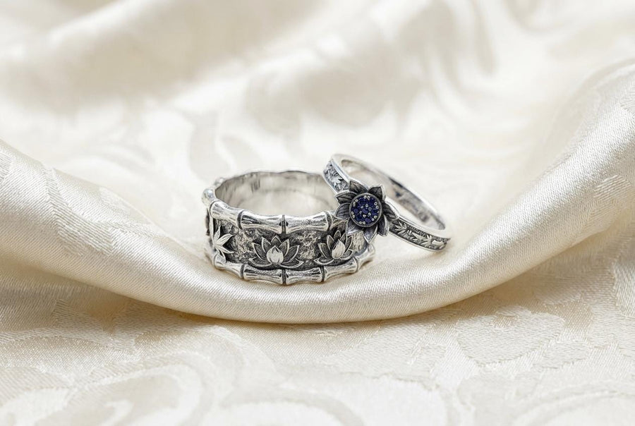 A close-up studio photograph of a matching 925 sterling silver botanical wedding ring set on a grey surface. The wide mens band features bamboo stalk edges and sculpted lotus flowers. The delicate womens engagement ring showcases a blooming lotus set with blue stones, highlighted by dark oxidation.