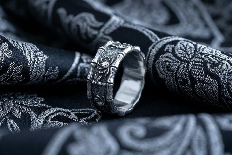 A close-up studio photograph of a matching 925 sterling silver botanical wedding ring set on a grey surface. The wide mens band features bamboo stalk edges and sculpted lotus flowers. The delicate womens engagement ring showcases a blooming lotus set with blue stones, highlighted by dark oxidation.