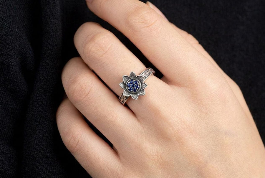 A close-up studio photograph of a matching 925 sterling silver botanical wedding ring set on a grey surface. The wide mens band features bamboo stalk edges and sculpted lotus flowers. The delicate womens engagement ring showcases a blooming lotus set with blue stones, highlighted by dark oxidation.