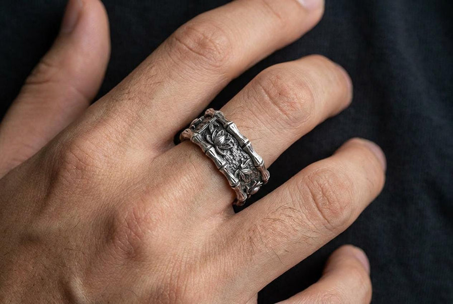 A close-up studio photograph of a matching 925 sterling silver botanical wedding ring set on a grey surface. The wide mens band features bamboo stalk edges and sculpted lotus flowers. The delicate womens engagement ring showcases a blooming lotus set with blue stones, highlighted by dark oxidation.