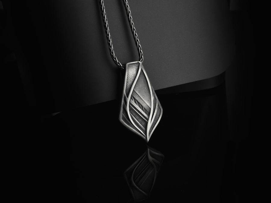 The pendant features a striking elongated shield silhouette, heavily detailed with precise micro-engraved lines and stippled textures that echo industrial elements. These darkened, oxidized recesses contrast brilliantly against the sharply polished, sweeping outer ridges. The multi-layered, armor-like plates give the piece incredible optical depth and a tactile, industrial feel.