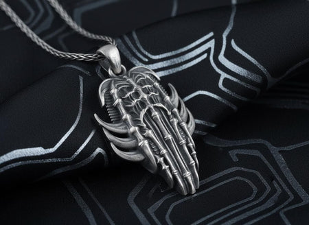Dark sci-fi silver skeleton pendant lying on black patterned fabric, showing intricate biomechanical ribs and an oxidized spine.