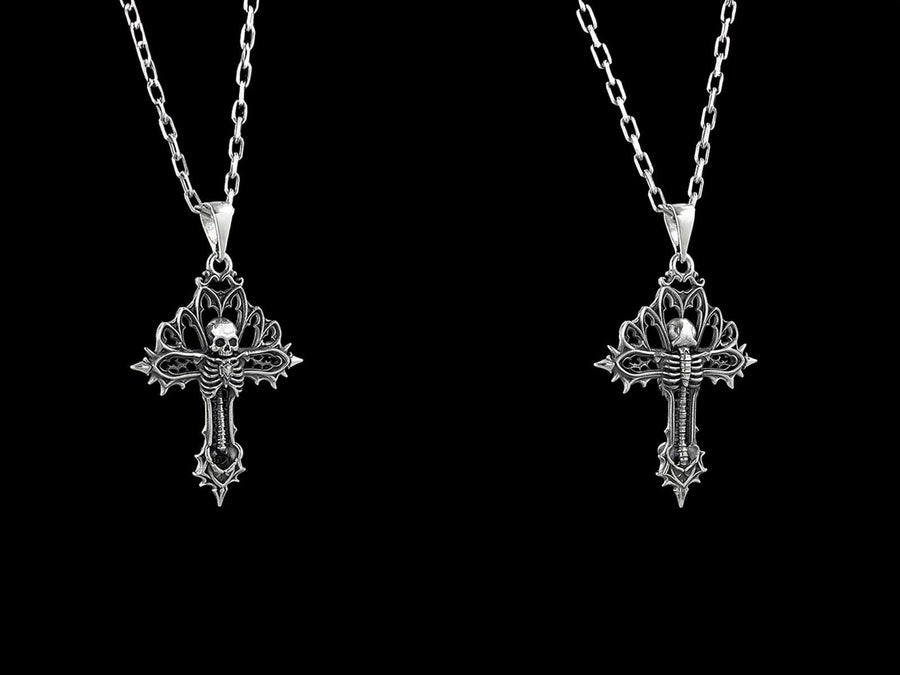 A close-up studio photograph of a 925 sterling silver gothic cross pendant resting on a light, finely textured surface. The core of the cross features a detailed skull, ribcage, and a spine extending down the bottom arm. The upper arms are crafted with openwork medieval tracery resembling cathedral windows. The recessed silver is heavily oxidized to a dark gray, contrasting sharply with the polished bone and architectural highlights.
