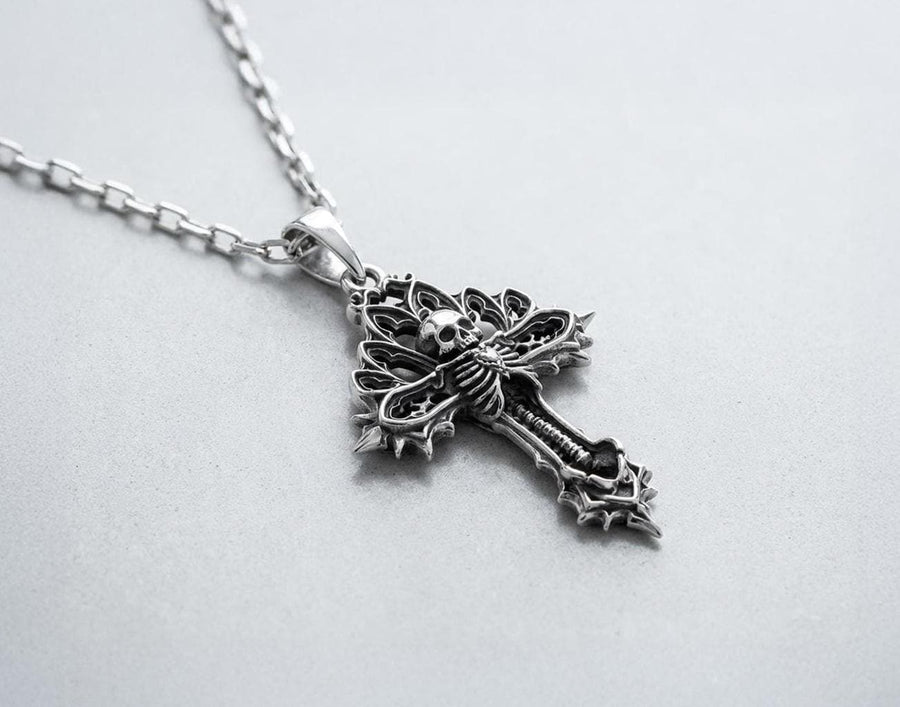 A close-up studio photograph of a 925 sterling silver gothic cross pendant resting on a light, finely textured surface. The core of the cross features a detailed skull, ribcage, and a spine extending down the bottom arm. The upper arms are crafted with openwork medieval tracery resembling cathedral windows. The recessed silver is heavily oxidized to a dark gray, contrasting sharply with the polished bone and architectural highlights.