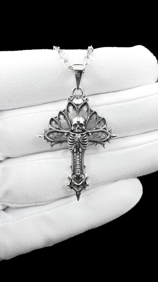 A close-up studio photograph of a 925 sterling silver gothic cross pendant resting on a light, finely textured surface. The core of the cross features a detailed skull, ribcage, and a spine extending down the bottom arm. The upper arms are crafted with openwork medieval tracery resembling cathedral windows. The recessed silver is heavily oxidized to a dark gray, contrasting sharply with the polished bone and architectural highlights.