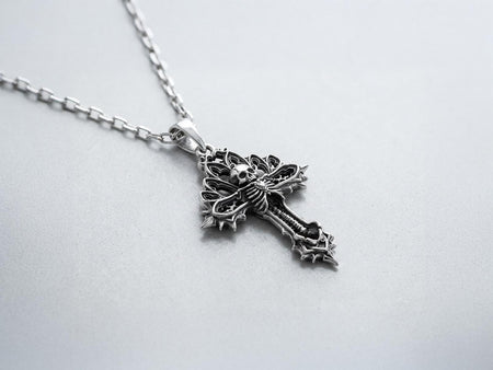 A close-up studio photograph of a 925 sterling silver gothic cross pendant resting on a light, finely textured surface. The core of the cross features a detailed skull, ribcage, and a spine extending down the bottom arm. The upper arms are crafted with openwork medieval tracery resembling cathedral windows. The recessed silver is heavily oxidized to a dark gray, contrasting sharply with the polished bone and architectural highlights.