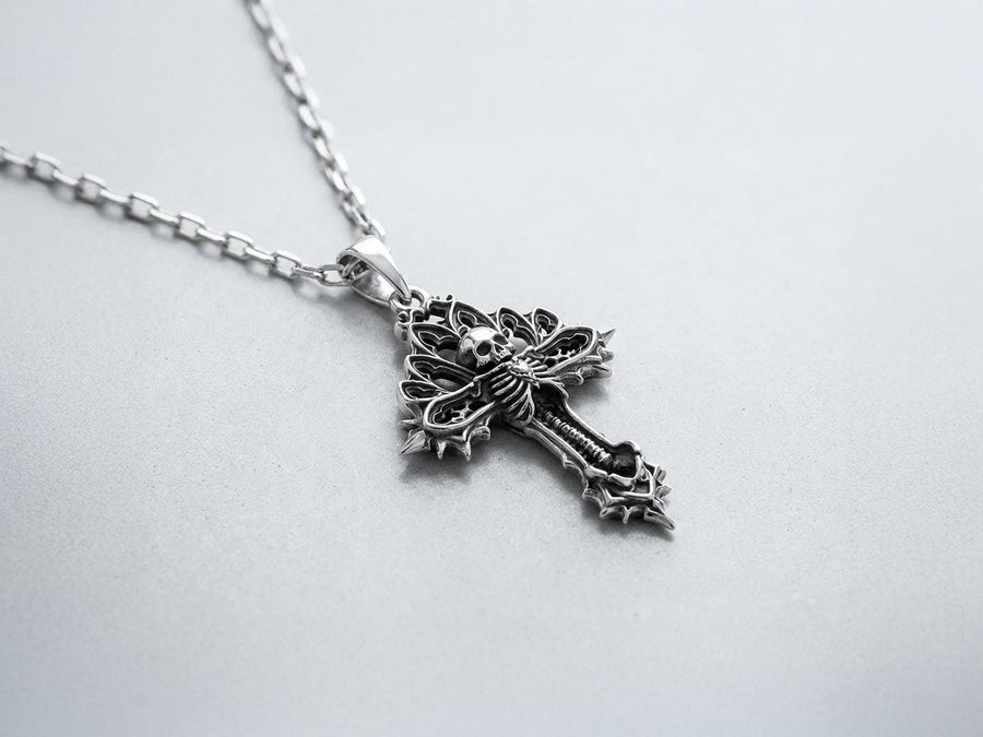 A close-up studio photograph of a 925 sterling silver gothic cross pendant resting on a light, finely textured surface. The core of the cross features a detailed skull, ribcage, and a spine extending down the bottom arm. The upper arms are crafted with openwork medieval tracery resembling cathedral windows. The recessed silver is heavily oxidized to a dark gray, contrasting sharply with the polished bone and architectural highlights.
