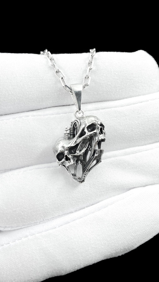 A side-profile view of the silver skull pendant showcases the breathtaking depth of the sculpted elements. The skulls appear stretched and elongated, morphing into each other. Dark oxidation heavily shadows the deeply recessed areas of the bone.