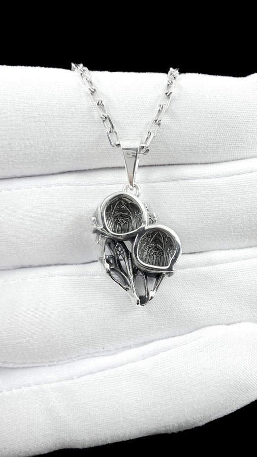 A side-profile view of the silver skull pendant showcases the breathtaking depth of the sculpted elements. The skulls appear stretched and elongated, morphing into each other. Dark oxidation heavily shadows the deeply recessed areas of the bone.