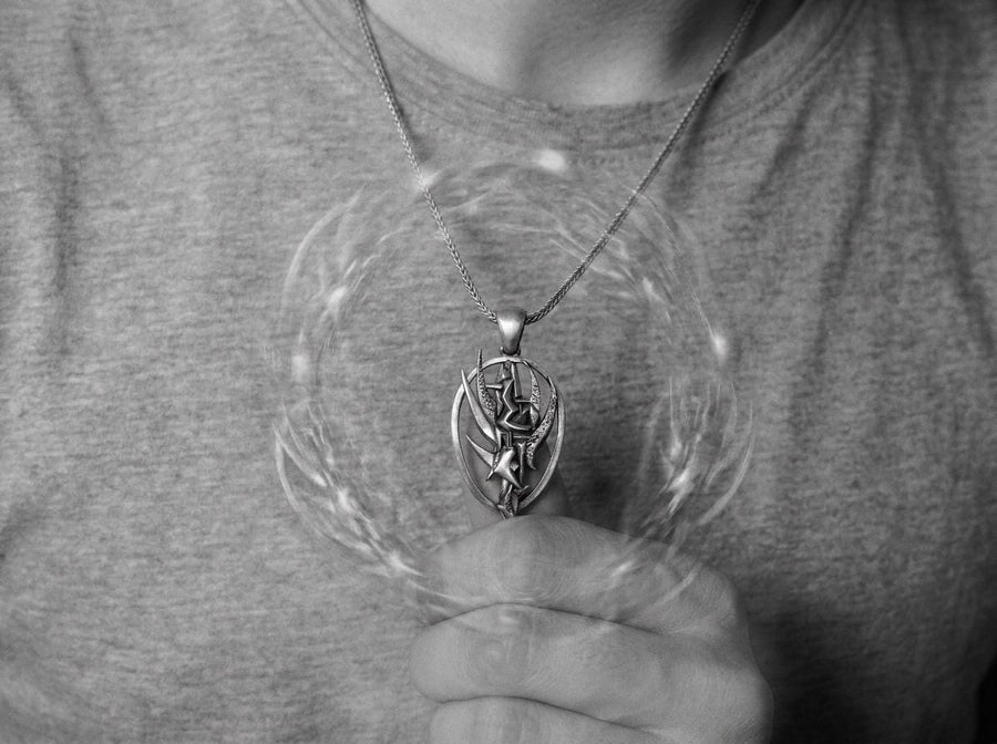 Silver dystopian techwear charm hanging on an anchor chain, emphasizing its rebellious asymmetrical design. A close-up studio photograph of a 925 sterling silver hieroglyph pendant resting on a dark patterned fabric. The avant-garde oval design features a sharp, geometric abstract symbol in the center, highlighted by a dark matte oxidized finish.
