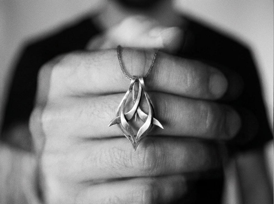 Solid sterling silver esoteric dagger drop charm held in hand, emphasizing the aggressive yet elegant lines. Close up of a man's fingers holding an abstract gothic spike amulet, highlighting the openwork silver design. Exceptional silversmithing is evident in this oxidized silver avant-garde necklace. The relief work perfectly balances the sharp lower points with smooth, sweeping interlaced upper curves. A bold modern statement piece for alte