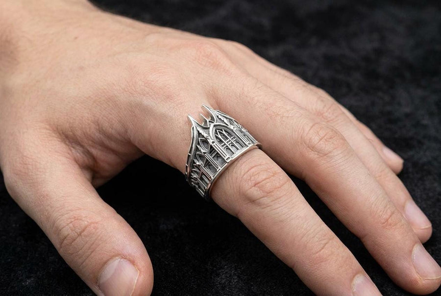 This solid 925 sterling silver gothic cathedral ring features soaring spires and intricately carved pointed arches. Resting on a grey surface, this oxidized silver band showcases detailed medieval tracery and classic stonework columns, perfect for a dark romance aesthetic. A striking 925 sterling silver architecture ring capturing the monumental scale of medieval sanctuaries. Bright lighting illuminates the polished silver gothic spires, contrast