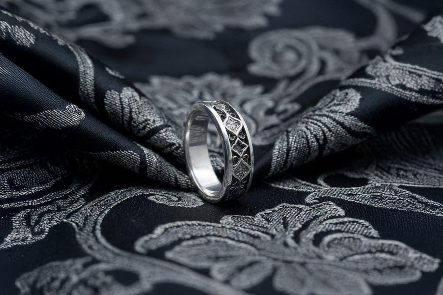 A close-up studio photograph of a matching 925 sterling silver gothic wedding ring set on a grey surface. The wide mens band and the delicate womens engagement ring both feature intricate medieval tracery and carved quatrefoils - highlighted by a dark oxidized background. This striking artisan bridal set perfectly captures the monumental scale of medieval architecture. Bright lighting illuminates the polished silver edges and the sparkling oval g