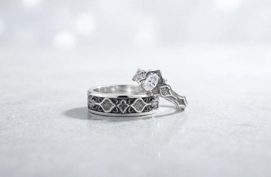 A close-up studio photograph of a matching 925 sterling silver gothic wedding ring set on a grey surface. The wide mens band and the delicate womens engagement ring both feature intricate medieval tracery and carved quatrefoils - highlighted by a dark oxidized background. This striking artisan bridal set perfectly captures the monumental scale of medieval architecture. Bright lighting illuminates the polished silver edges and the sparkling oval g