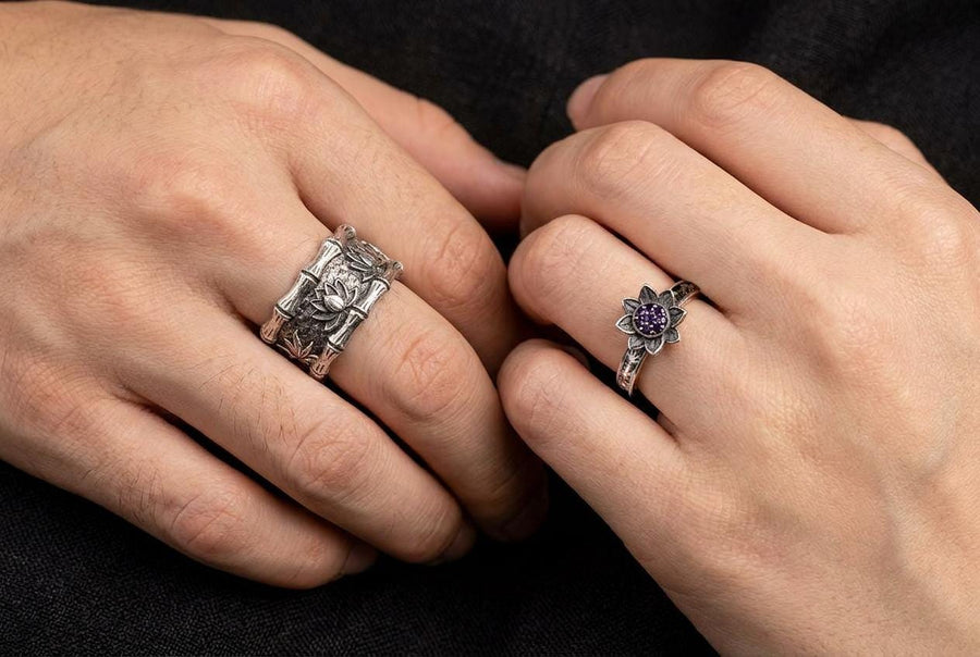 A close-up studio photograph of a matching 925 sterling silver botanical wedding ring set on a grey surface. The wide mens band features bamboo stalk edges and sculpted lotus flowers. The delicate womens engagement ring showcases a blooming lotus set with blue stones, highlighted by dark oxidation.