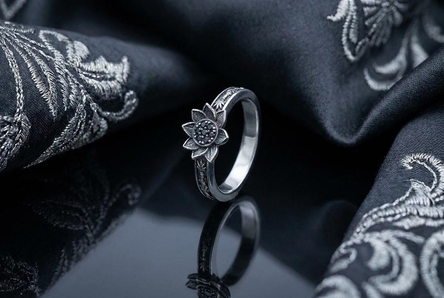 A close-up studio photograph of a matching 925 sterling silver botanical wedding ring set on a grey surface. The wide mens band features bamboo stalk edges and sculpted lotus flowers. The delicate womens engagement ring showcases a blooming lotus set with blue stones, highlighted by dark oxidation.