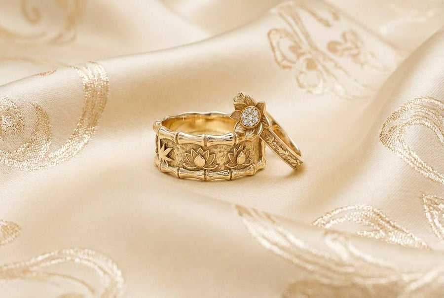 A close-up studio photograph of a matching 925 sterling silver botanical wedding ring set on a grey surface. The wide mens band features bamboo stalk edges and sculpted lotus flowers. The delicate womens engagement ring showcases a blooming lotus set with blue stones, highlighted by dark oxidation.