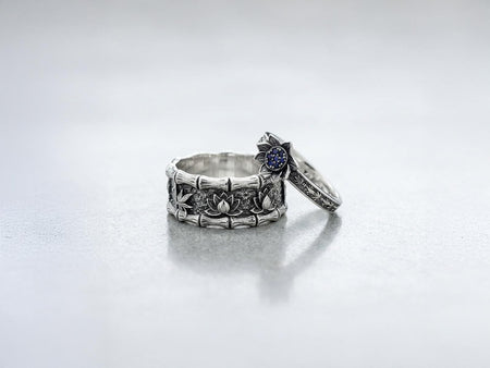 A close-up studio photograph of a matching 925 sterling silver botanical wedding ring set on a grey surface. The wide mens band features bamboo stalk edges and sculpted lotus flowers. The delicate womens engagement ring showcases a blooming lotus set with blue stones, highlighted by dark oxidation.