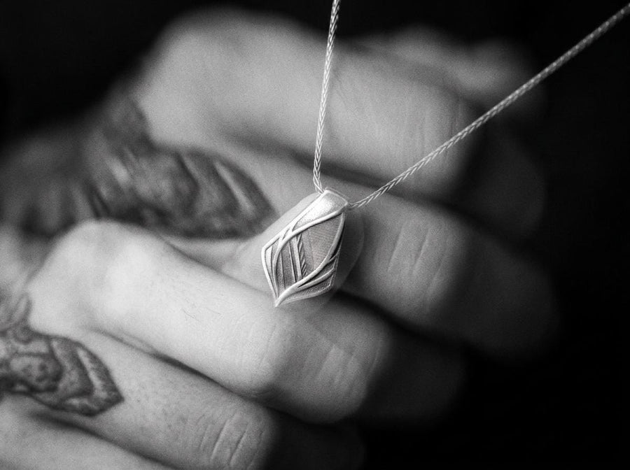 The pendant features a striking elongated shield silhouette, heavily detailed with precise micro-engraved lines and stippled textures that echo industrial elements. These darkened, oxidized recesses contrast brilliantly against the sharply polished, sweeping outer ridges. The multi-layered, armor-like plates give the piece incredible optical depth and a tactile, industrial feel.
