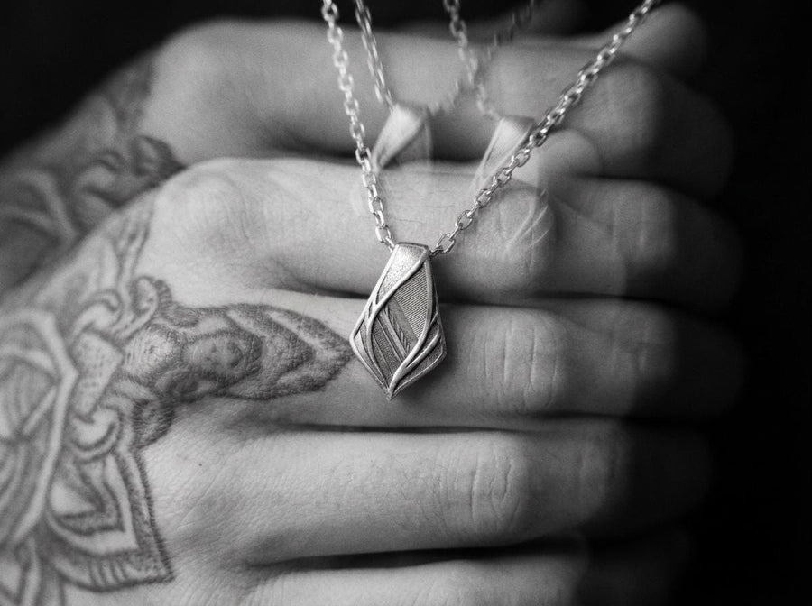The pendant features a striking elongated shield silhouette, heavily detailed with precise micro-engraved lines and stippled textures that echo industrial elements. These darkened, oxidized recesses contrast brilliantly against the sharply polished, sweeping outer ridges. The multi-layered, armor-like plates give the piece incredible optical depth and a tactile, industrial feel.