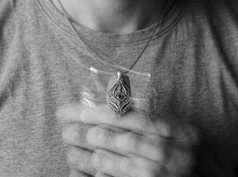 Unique artisan crafted silver armor necklace for men, fantasy sci-fi jewelry, A close-up studio photograph of a 925 sterling silver shield pendant resting on a dark reflective surface. The avant-garde design features geometric plating and a central circular core, highlighted by a dark matte oxidized finish. This striking artisan pendant captures the essence of futuristic armor.