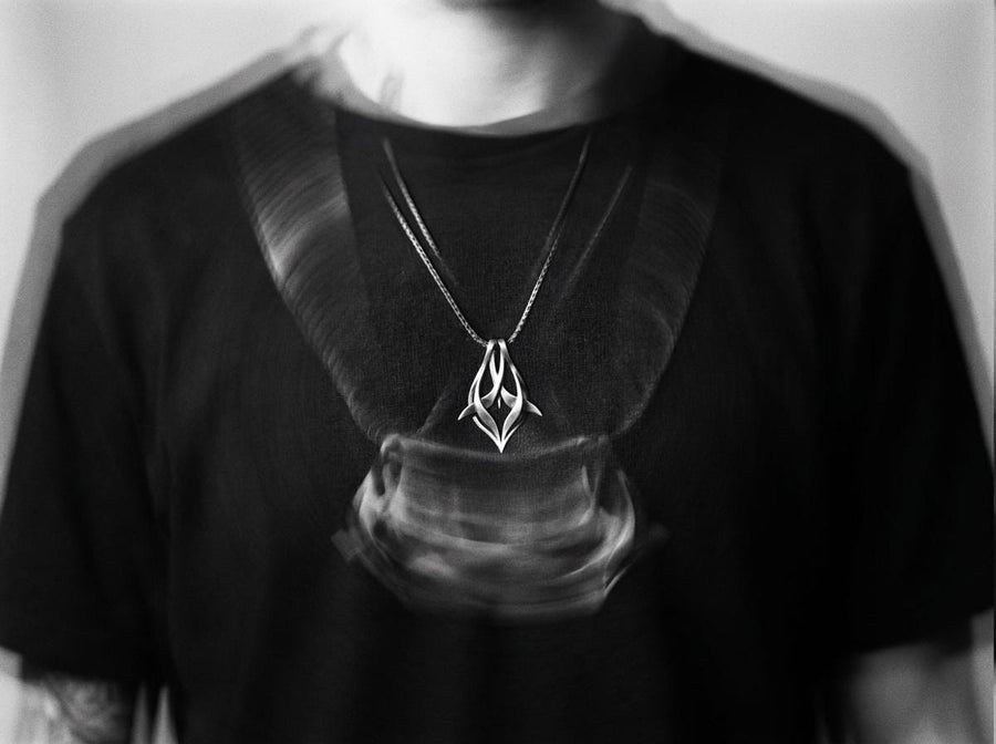 Solid sterling silver esoteric dagger drop charm held in hand, emphasizing the aggressive yet elegant lines. Close up of a man's fingers holding an abstract gothic spike amulet, highlighting the openwork silver design. Exceptional silversmithing is evident in this oxidized silver avant-garde necklace. The relief work perfectly balances the sharp lower points with smooth, sweeping interlaced upper curves. A bold modern statement piece for alte